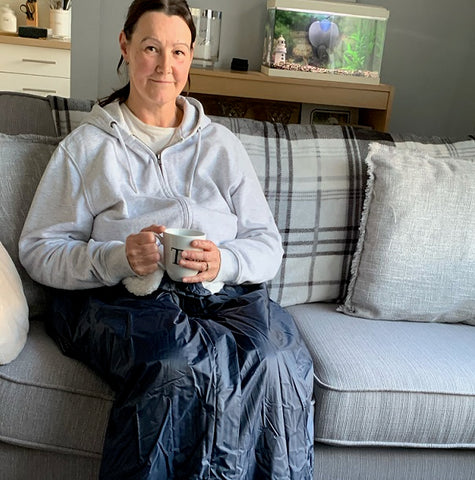 The fleece lined leg cosy comes in a choice of cream or blue fleece lining. It has a waterproof outer. Here, a woman is wearing one whilst sat on the couch, drinking a cup of tea.