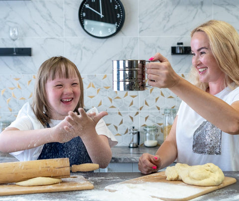 This is a photograph of a mother and daughter, who has Down's syndrome, baking together. They are both smiling, as the mother sprinkles powder on the worktop.