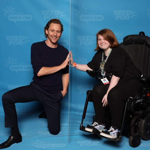 Ami is sitting in her new wheelchair. Tom Hiddleston is to the left of Ami – a sheet of glass separates them (covid precaution) and each person has one hand against the glass