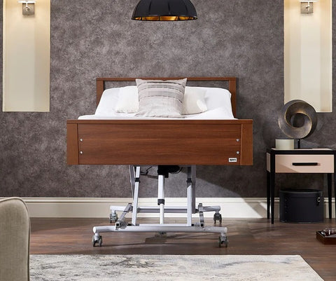 This is a picture of the Opera Low Footboard Bed when it is raised up to allow for a carer to tend to the person. It has a dark walnut footboard and silver structures underneath.