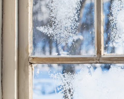 A rustic wooden sash window with ice patterns on the glass. There is a wintery landscape outside, and everything is covered in snow.
