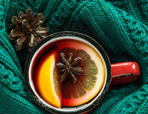 A photo of a red ceramic mug full of mulled wine resting on a green blanket. There are two citrus wedges and star anise in the mulled drink, a spice associated with winter in the UK.