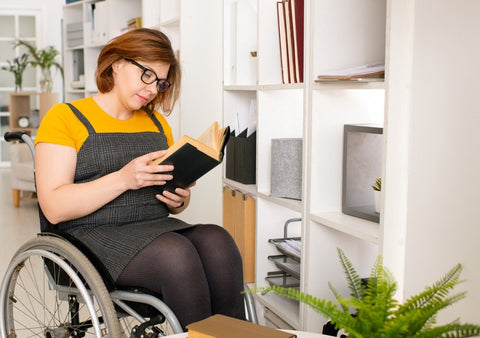 An image of a young woman using a wheelchair. She is reading a book and has a variety of houseplants around.