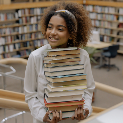 A woman with natural hair holding a stack of books in a library