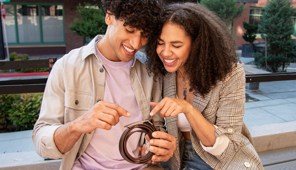 6th Anniversary Iron Gift Ideas like a custom belt buckle are perfect for your husband or wife. Image of couple looking at new custom belt buckle at an outdoor cafe, sitting close together and smiling.