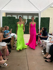 Two models walking down the runway. One is wearing a neon yellow sheath dress, the other is wearing a neon pink sheath dress. They both have a very serious look on their faces.