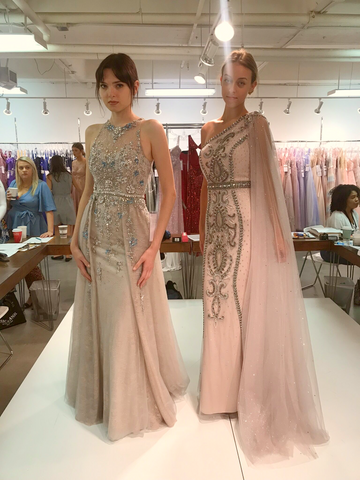 two models on a runway. One is wearing a blush sheath gown with lots of beading. The other is wearing a one shoulder blush gown with beading down the front and a cape coming off the one shoulder.
