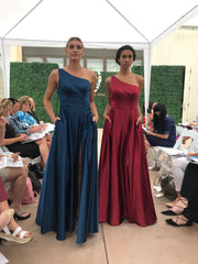 Two models walking down the runway. One is wearing a one shoulder, navy blue prom dress. The other is wearing a one shoulder, wine colored prom dress.