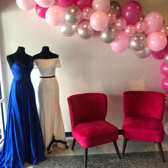 Inside XO's storefront. There are two hot pink velvet chairs next to two prom gowns. One is a Royal Blue A line Gown and the other is a white off the shoulder two piece sheath gown. There is a Hot Pink, Silver, and Light Pink balloon arch on the right going over the top of the chairs and dresses.