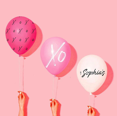 three balloons in descending height being held by three hands. The tallest one is hot pink and has black Xs and Os in it. The middle balloon is light pink and says XO very big in white. The shortest one is white and says By Sophia's in black. They are on a light pink backdrop.