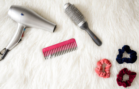 Hairdryer, comb, barrelbrush, and scrunchies laying out flat on a white fur surface. 