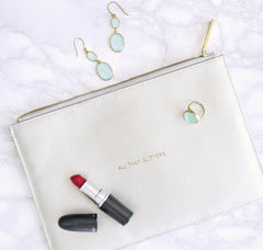 A white clutch that is laying on a marble background. It has a tube of red lipstick and teal earrings laying on top of it.