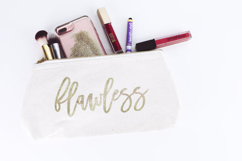 A white clutch with gold writing that says Flawless on it is laying on a white background. It has makeup brushes, lipstick, an iphone, and a pen sticking out of the top.