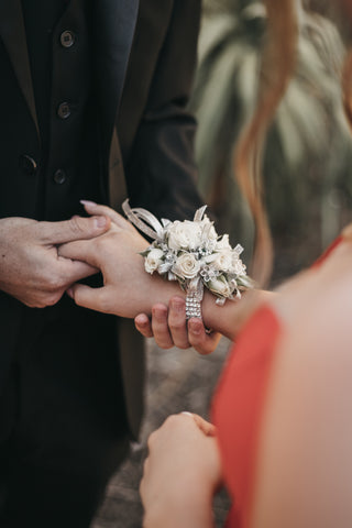 The view is over the shoulder of a prom girl. Her date is holding her hand a putting a corsage on her wrist. The flowers are white roses.