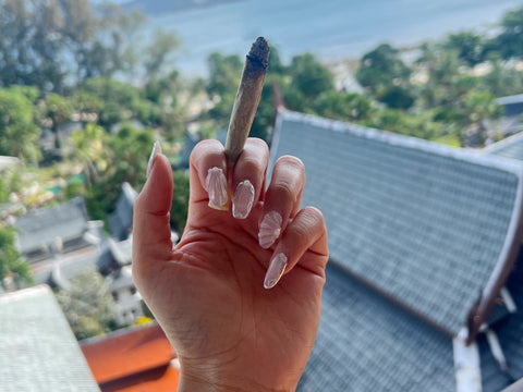 Woman's hand with mermaid shell acrylic nail art holding a big joint in Phuket, Thailand in front of a beach view.
