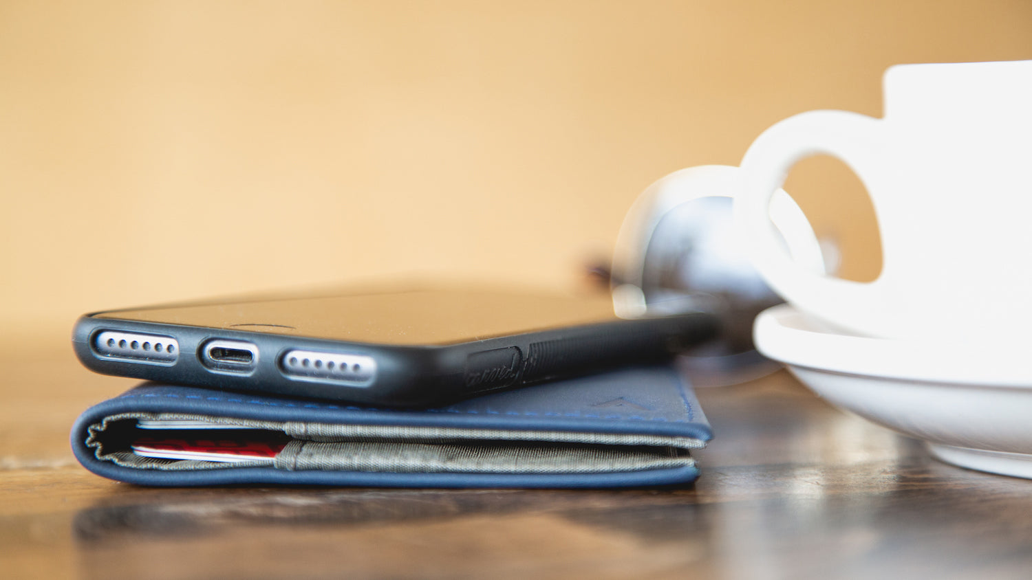 Das iPhone liegt auf einer mitternachtsblauen Allett-Originalbrieftasche aus Leder auf einer Tischplatte neben einer Tasse Kaffee auf einer Untertasse