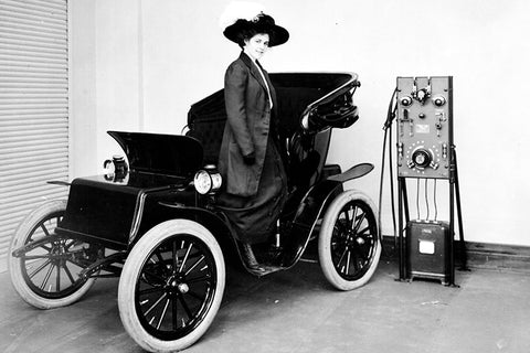 A woman stands inside a early 1900's electric car automobile which is being charged by a mercury arc rectifier electric car charging station.