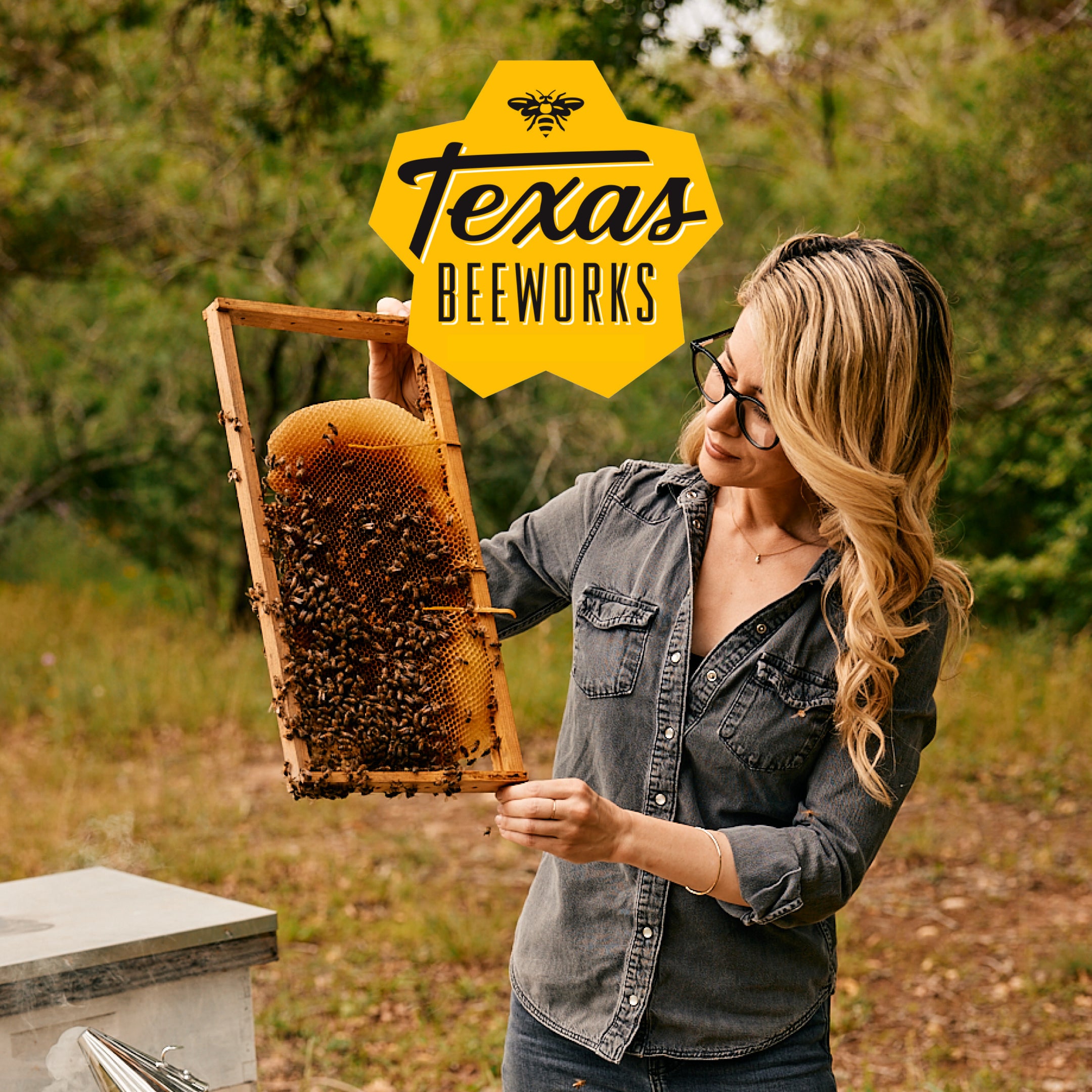 Woman inspecting a honeycomb frame from a hive, with Texas Beeworks logo.