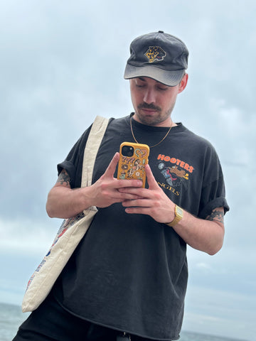 Artist Cam Miller looks down at the phone in his hands, which is encased in the Pela Honey Miller West phone case he designed. He is wearing a black cap, a black Hooters Angels t-shirt, and a canvas bag over his shoulder. The background is a cloudy sky.