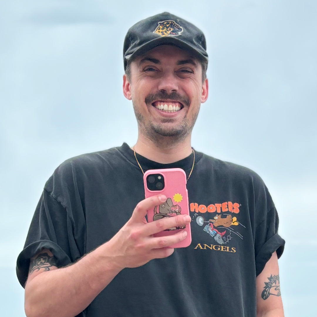 Smiling man wearing a cap and t-shirt taking a selfie with a phone.