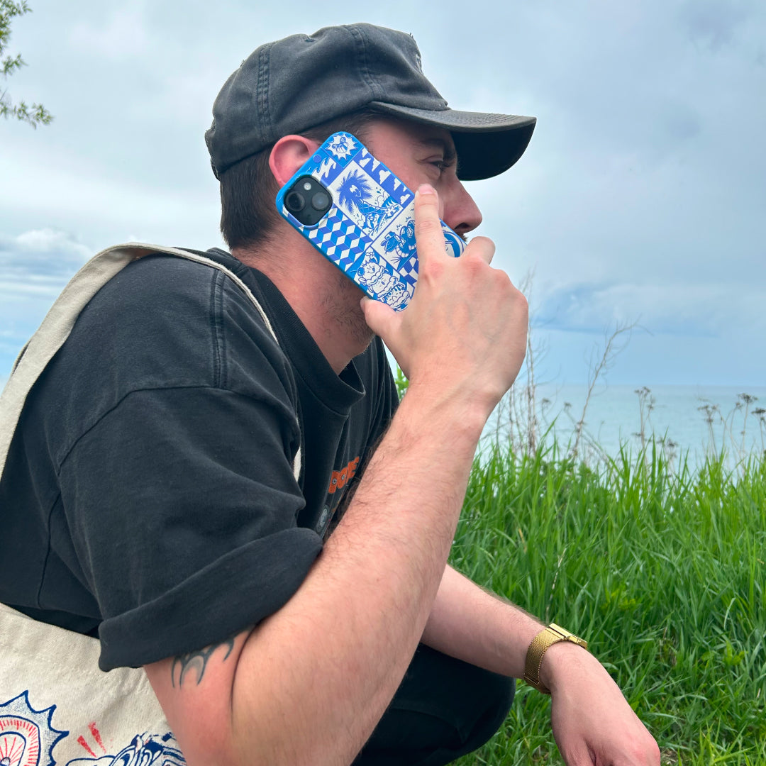 Person sitting outdoors on grass, talking on a blue patterned phone, with water and sky in the background.