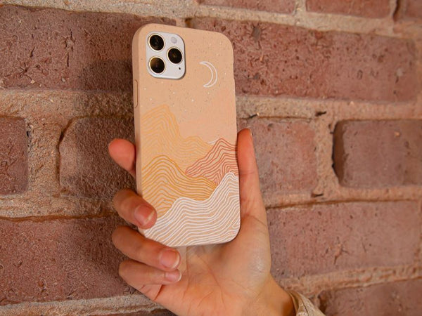 A phone in a seashell pink peaks Pela case held against a brick wall