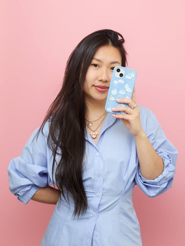 Janine from Yeunglove holding a phone case featuring the 'Fluffy Flower on Powder Blue' design, a collaboration with Pela. The background is a soft, powder blue color, highlighting the intricate and playful design of the case. Janine is smiling, showcasing the unique artwork with pride.