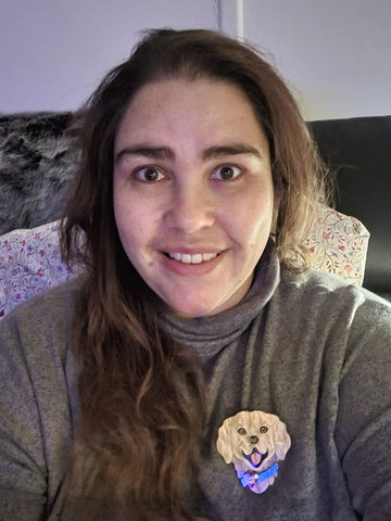 Image Description: Nay, a First Nations woman, wearing a grey turtleneck, with long, wavy, brown hair smiles widely. She wears a Limited Edition She Loves Blooms x Poly Paige Service Dog brooch, the brooch is the head and shoulders of a smiling, golden furred dog, with a blue collar. On the collar is a red heart and the words Love and Loyalty printed on it. 