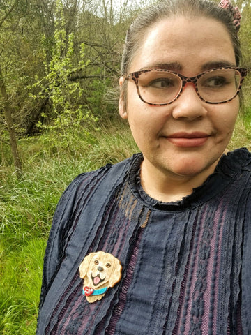 Image description: Nay, a First Nations woman, wearing a purple top, animal print glasses and she looks off frame in front of thick bushland. She wears a Limited Edition She Loves Blooms x Poly Paige Service Dog brooch, the brooch is the head and shoulders of a smiling, golden furred dog, with a blue collar. On the collar is a red heart and the words Love and Loyalty printed on it. 