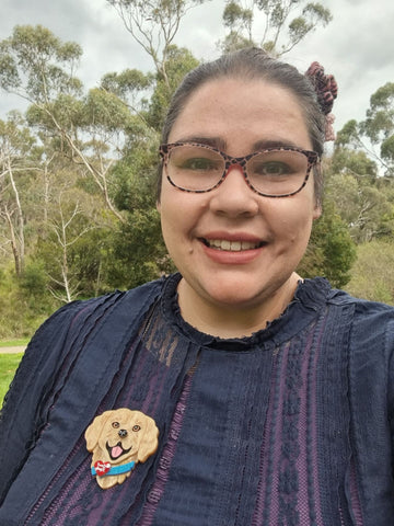 Image description: Nay, a First Nations woman, wearing a purple top, animal print glasses and smiling widely sits in front of thick bushland. She wears a Limited Edition She Loves Blooms x Poly Paige Service Dog brooch, the brooch is the head and shoulders of a smiling, golden furred dog, with a blue collar. On the collar is a red heart and the words Love and Loyalty printed on it. 