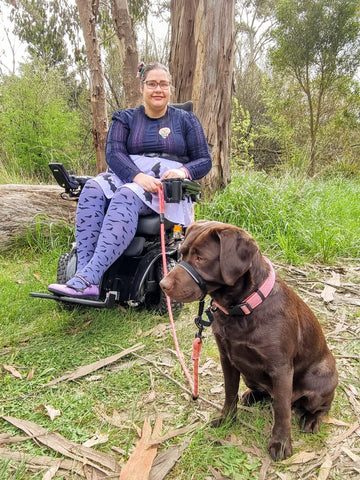 Image description: Nay, a First Nations woman wearing a purple outfit and a very cute She Loves Blooms x Poly Paige Service Dog brooch, sits in a big black wheelchair out in the bush. Luna, a chocolate brown Labrador sits in front of her wearing a halter, lead, and collar. 