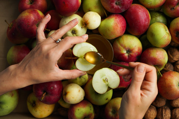 two hands holding red and green apple slices with spoon of honey