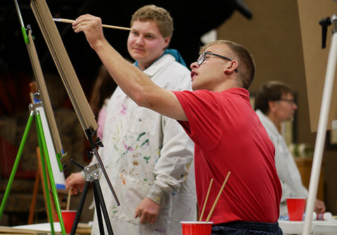 One adult man paints a fine art canvas while standing at an easel with another adult man looking on with approval of the work.