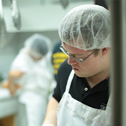 Image of Nolan Stillwell, a young man with Down syndrome in
                a commercial kitchen wearing a white apron, navy shirt and a
                hairnet.