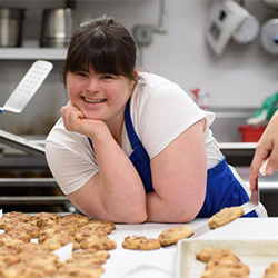 Image of Collettey, a woman with Down syndrome, in a
                commercial kitchen wearing a blue apron smiling with some
                cookies.