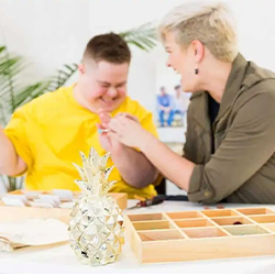 Image of Margrit Co. founder, Maggie with her brother who
                was born with Down syndrome. They are sitting at a desk making
                jewellry.