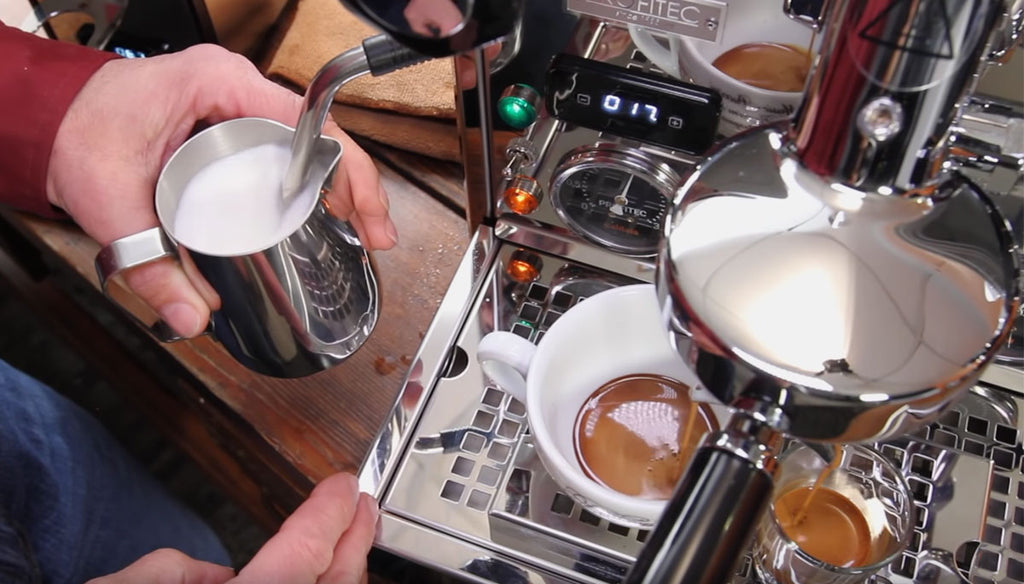 Milk inside of a stainless steel carafe is steamed with the Pro 700's steam wand inside of it. Two shots are being pulled as well, one in a cappuccino cup, and the other in a shot glass.