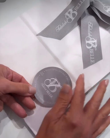 A woman's hands apply a logo sticker to a gift bag for Talulah Belle. A gray ribbon is tied in a bow on the white gift bag with the brand logo.