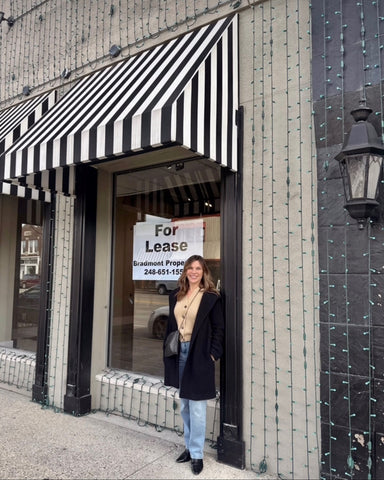 Dark blonde haired woman wearing a black jacket standing in front of a black and white striped awning storefront of the futureTalulah Belle Boutique in Rochester Michigan. Sign in the window reads 'For Lease'.