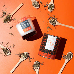 White Tea & Bergamot Luxury scented candle from Lower Lodge Candles Colour Pop! collection pictured with its cardboard tube packaging against an orange background with wooden spoon with tea leaves on them