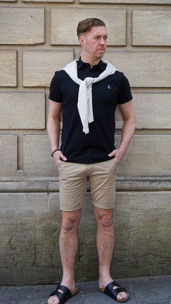 Model wearing black polo shirt, sand chino shorts, and sandals. 