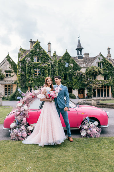 Couple outside venue face camera. Groom is wearing the Wells suit. Bride is wearing a pale pink dress which matches the hot pink car behind them.