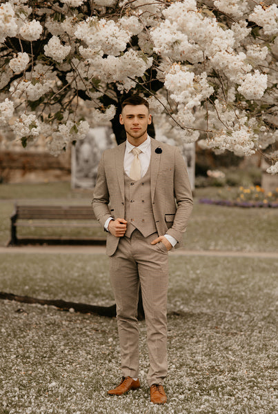 Cream suit for wedding worn by groom who stands underneath a blossoming tree.