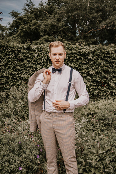 Model wears the Elwood suit with the jacket over his shoulder, plus a suave owl white shirt, black bow tie, and black braces. He is outside amongst greenery.