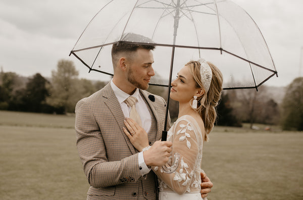 Groom wears cream groom's suit standing with bride under umbrella.