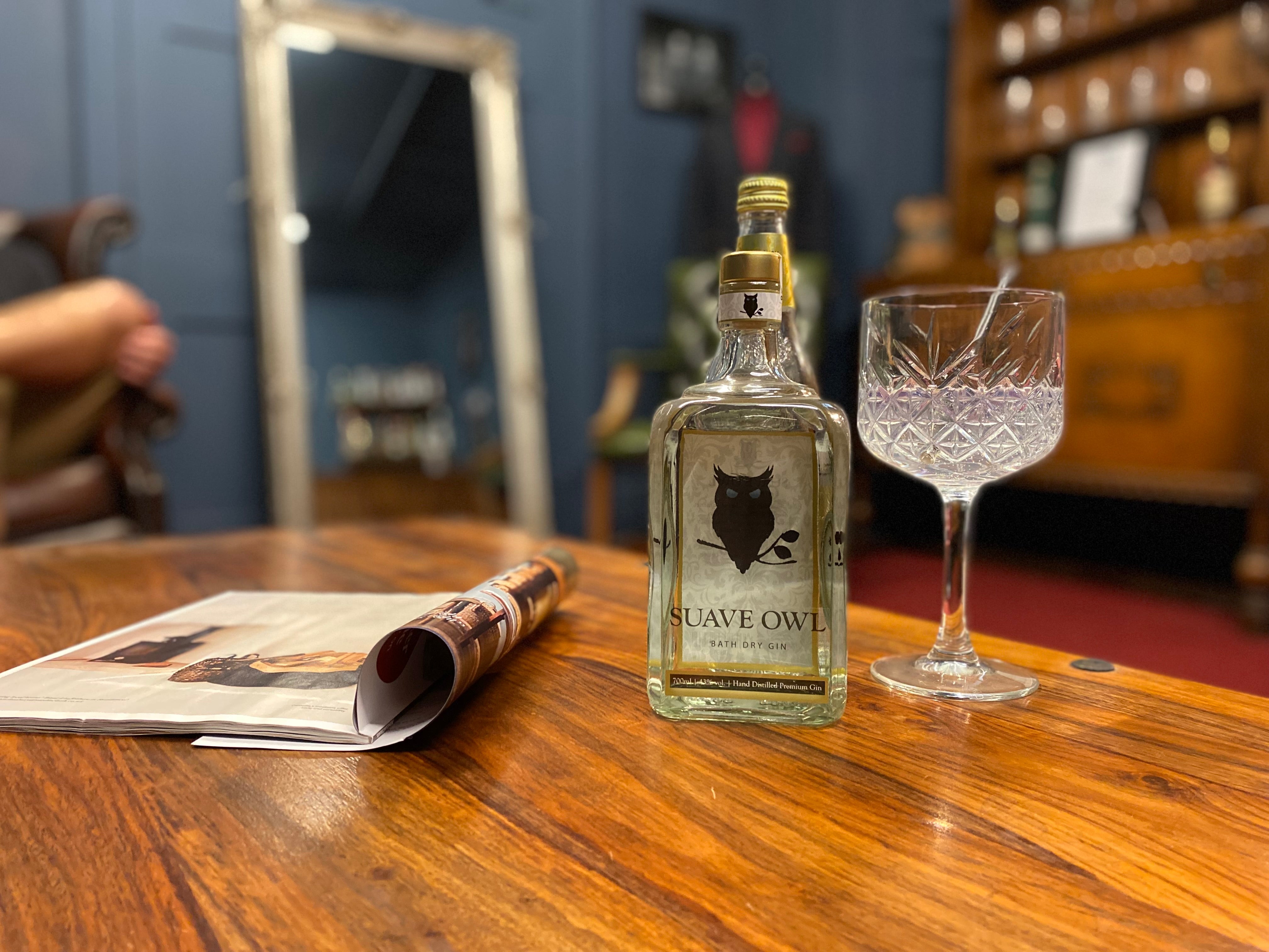 SUAVE OWL gin sits on a wooden table. There is a crystal glass with a paper straw. The room is finely decorated but slightly blurred in the background. 