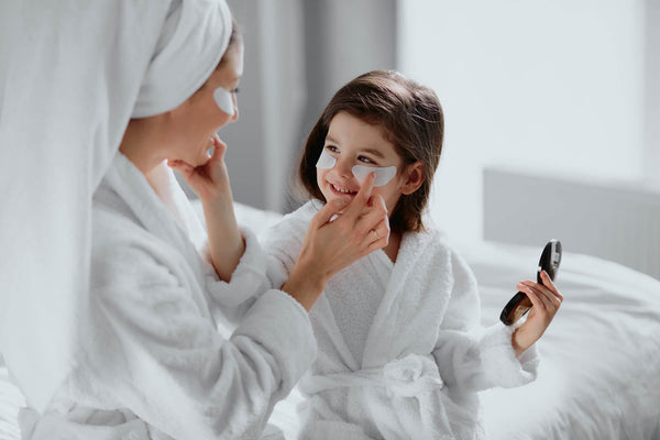 Cute mother and daughter sit together on bed in beauty masks patches at home