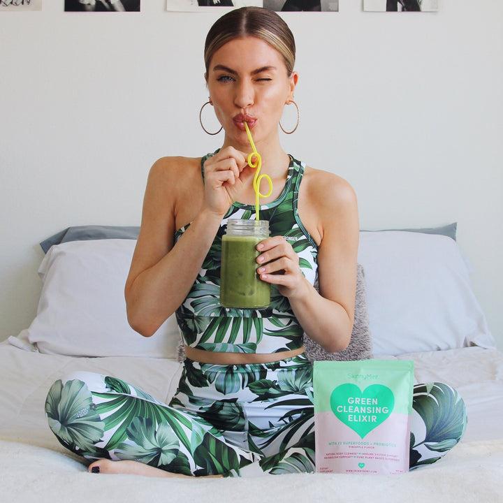 A model drinking Green Cleansing Elixir while sitting on her bed. 