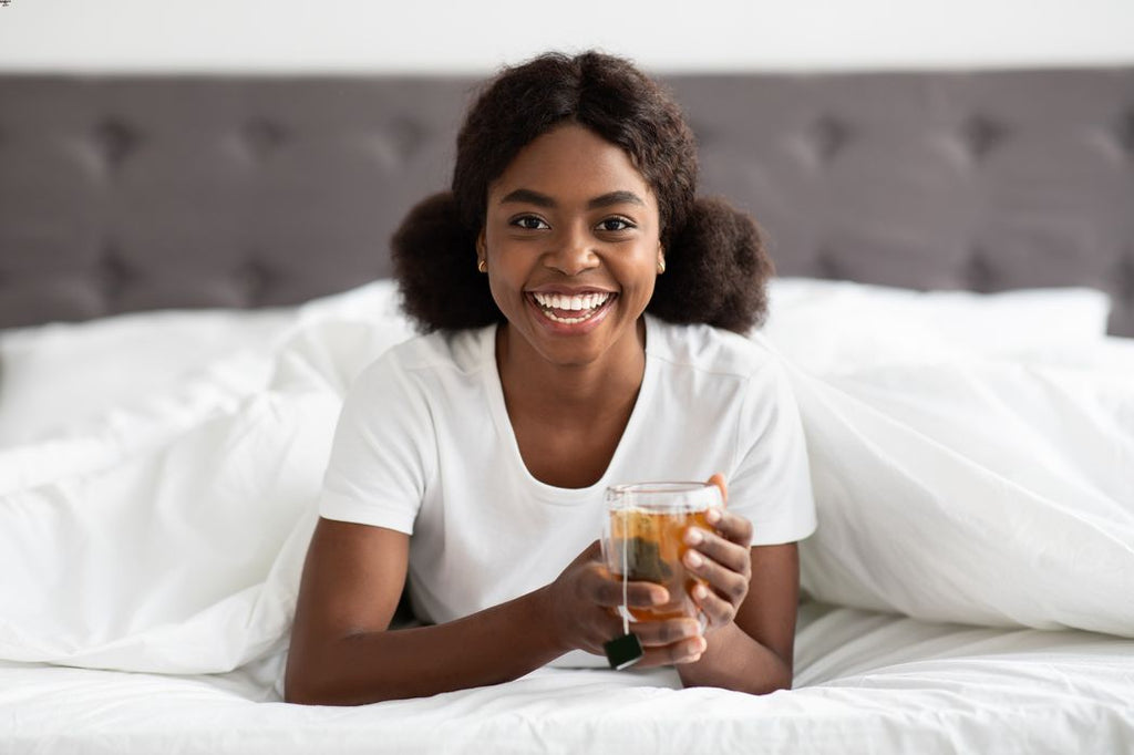 A smiling Black woman drinks green tea in bed.