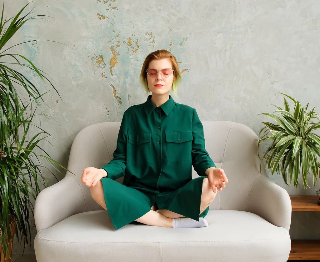 A woman sitting on a couch and meditating to reduce stress and her tendency to overeat.
